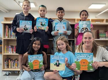 Six students and 1 Adult holding up books in front of them in the library.