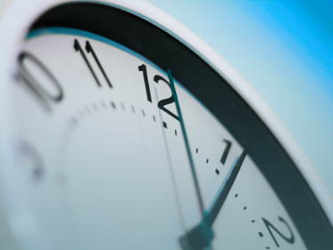 Close-up of an analog clock face showing the numbers 11, 12, 1, and 2, with the clock hands visible against a soft blue background.